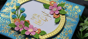 hand made teal card with tiny gold branches inlaid, a gold framed center sentiment surrounded by pink and orange flowers created with new card making supplies from Spellbinders