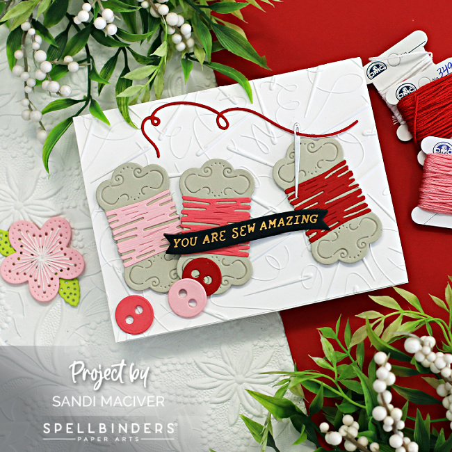 white embossed card base with three colors of pink embroidery floss skeins across the front, with pink and red buttons and a threaded needle. Created with new card making products from Spellbinders 