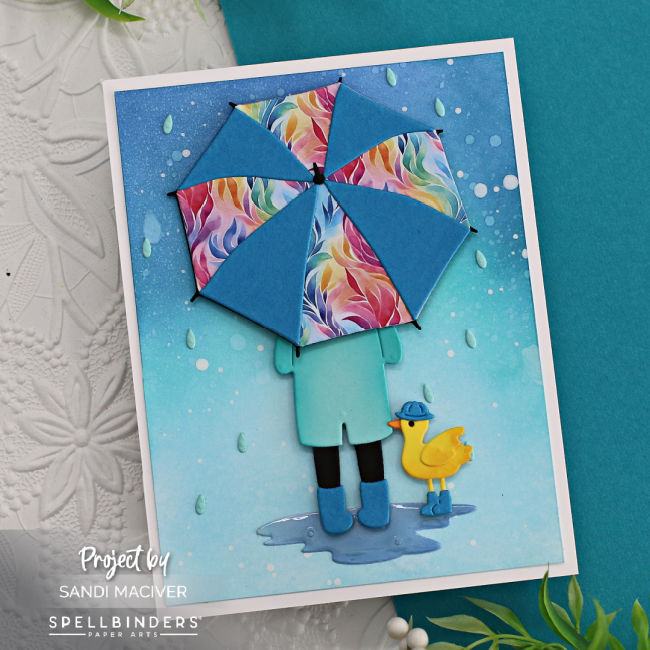 hand made card with a ombre blue ink blended background splattered to look like rain, a person holding a bright umbrella and a little yellow duck wearing blue rain boots created with card making dies from Spellbinders 