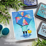 hand made card with a ombre blue ink blended background splattered to look like rain, a person holding a bright umbrella and a little yellow duck wearing blue rain boots created with card making dies from Spellbinders