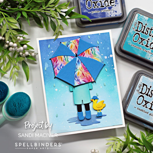 hand made card with a ombre blue ink blended background splattered to look like rain, a person holding a bright umbrella and a little yellow duck wearing blue rain boots created with card making dies from Spellbinders 