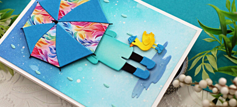 hand made card with a ombre blue ink blended background splattered to look like rain, a person holding a bright umbrella and a little yellow duck wearing blue rain boots created with card making dies from Spellbinders