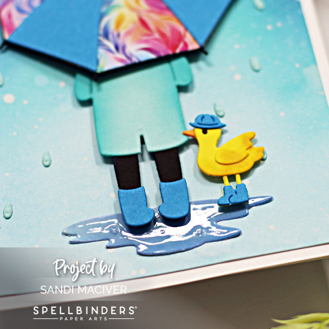 hand made card with a ombre blue ink blended background splattered to look like rain, a person holding a bright umbrella and a little yellow duck wearing blue rain boots created with card making dies from Spellbinders 