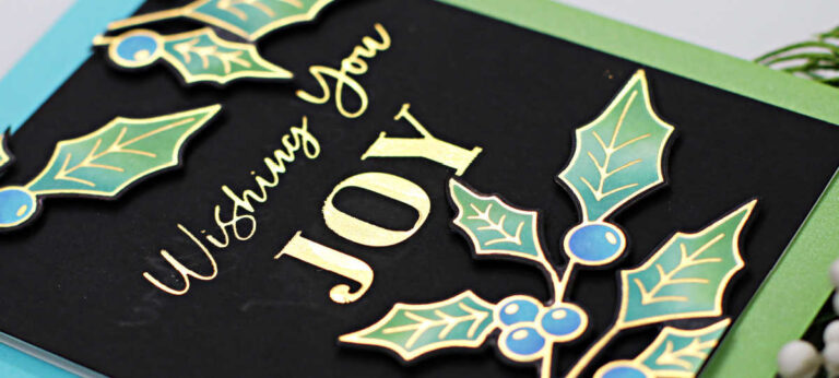 hand made christmas card with a black background, shinny gold sentiment and gold outlined holly with blue berries and three tone green leaves created with new card making products from Spellbinders