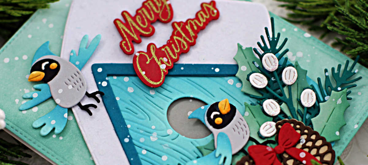 hand made christmas card with a teal snowcapped birdhouse, lots of green and white foliage, two blue and gray birds and red Merry Christmas sentiment created with new card making products from Spellbinders