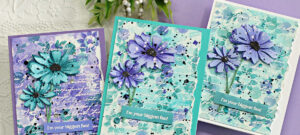 three hand made mixed media cards in teal and purple with stenciling and splatter and purple and teal die cut flowers