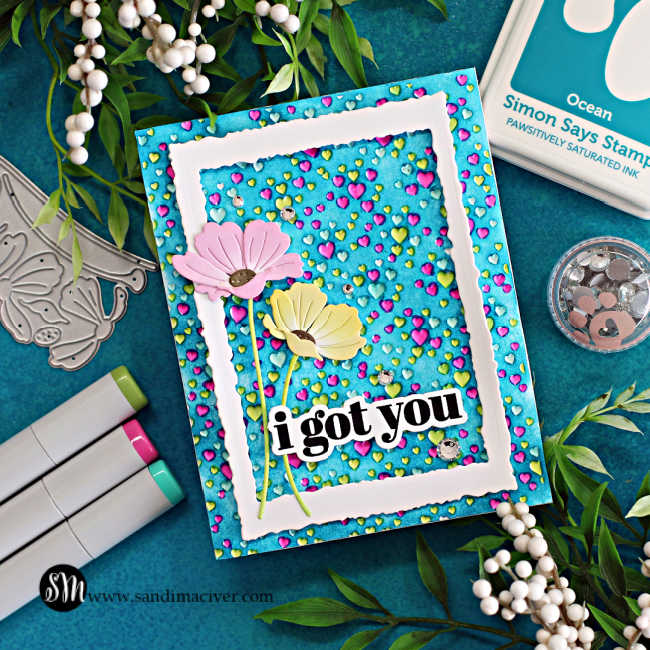 hand made card with a embossed heart background, the hearts colored in blue, green and pink, with a blue background, a white frame overlay and a pink and yellow die cut poppy on the left side 