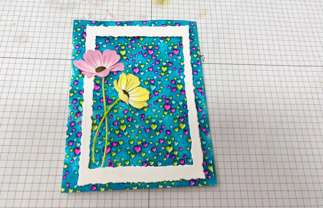 hand made card with a embossed heart background, the hearts colored in blue, green and pink, with a blue background, a white frame overlay and a pink and yellow die cut poppy on the left side 