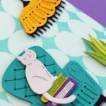 a retro feel hand made card with a two tone teal background, a teal stitched chair with a gray cat sitting atop some books with a yellow lamp over a plant in a purple pot