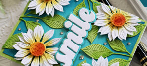 Handmade floral card featuring die-cut white sunflowers with yellow and orange centers, created with Simon Says Stamp Jaunty Sunflower dies. The flowers are layered over a teal background with dimensional green die-cut leaves and accented with scattered gems. A bold white “smile” sentiment die cut adds a cheerful touch. Perfect for summer-themed or uplifting handmade cards.