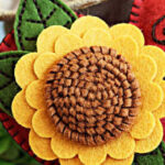 Handmade felt heart ornament featuring a layered sunflower with textured brown center, yellow petals, and green stitched leaves, created using Spellbinders dies. Embellished with brown embroidery swirl details on a rest felt heart, perfect for fall or farmhouse-style DIY craft, tiered tray display, and cardmaking projects