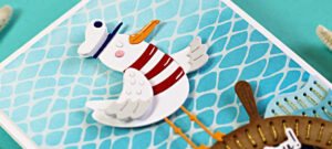 A hand-made card with a die-cut seagull dressed as a ship captain, standing on a stitched helm with a Glimmer foiled net background