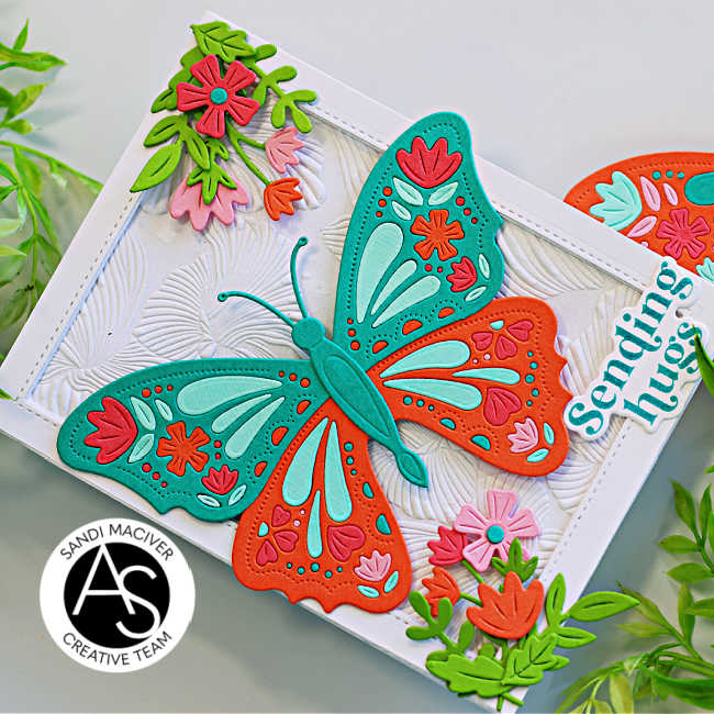 Handmade greeting card featuring a colorful die-cut butterfly design in teal, orange, pink, and aqua, accented with floral die cuts and embossed background texture. Sentiment reads “Sending hugs.” Perfect for spring or summer cardmaking inspiration using Alex Syberia Designs Butterfly Bliss collection.