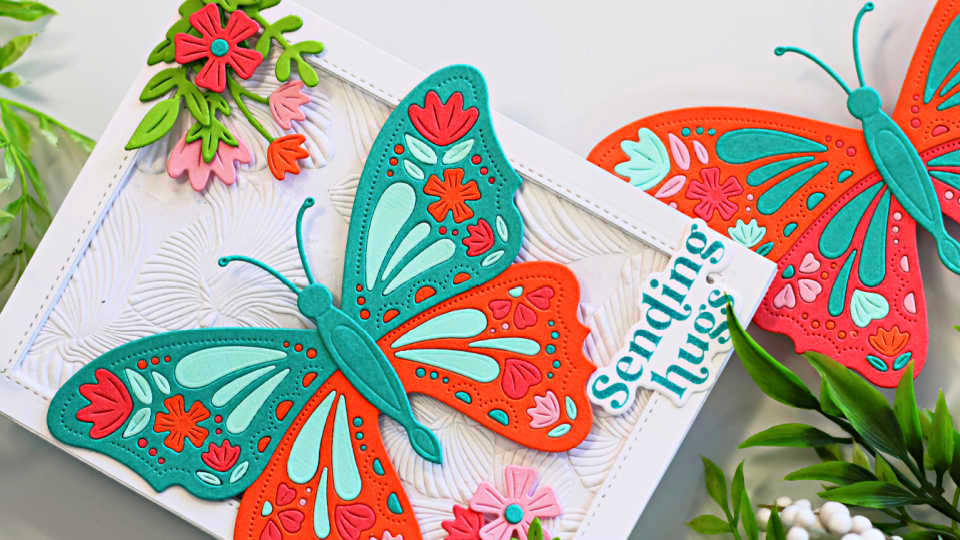 Handmade greeting card featuring a colorful die-cut butterfly design in teal, orange, pink, and aqua, accented with floral die cuts and embossed background texture. Sentiment reads “Sending hugs.” Perfect for spring or summer cardmaking inspiration using Alex Syberia Designs Butterfly Bliss collection.