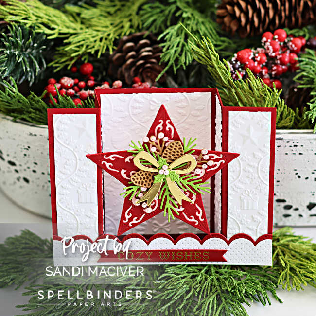 Handmade Christmas bridge card featuring a bold red dimensional star decorated with gold ribbon, pinecones, green sprigs, and white florals. The background includes embossed white panels with gift motifs and a pierced white scalloped bridge with a red 'Cozy Wishes' sentiment. Created by Sandi MacIver using Spellbinders dies.