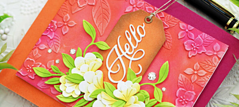 Handmade floral card created with Simon Says Stamp Happy Camper release products. Features an embossed pink and coral background with leafy floral designs, layered white and yellow die-cut flowers with bright green leaves, and a brown ink-blended tag with a white die-cut “Hello” sentiment. Embellished with sparkling clear gems for added dimension and shine.