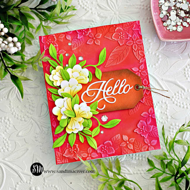 Handmade floral card created with Simon Says Stamp Happy Camper release products. Features an embossed pink and coral background with leafy floral designs, layered white and yellow die-cut flowers with bright green leaves, and a brown ink-blended tag with a white die-cut “Hello” sentiment. Embellished with sparkling clear gems for added dimension and shine.