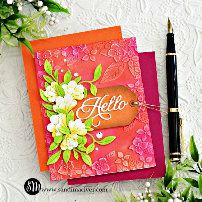 Handmade floral card created with Simon Says Stamp Happy Camper release products. Features an embossed pink and coral background with leafy floral designs, layered white and yellow die-cut flowers with bright green leaves, and a brown ink-blended tag with a white die-cut “Hello” sentiment. Embellished with sparkling clear gems for added dimension and shine.