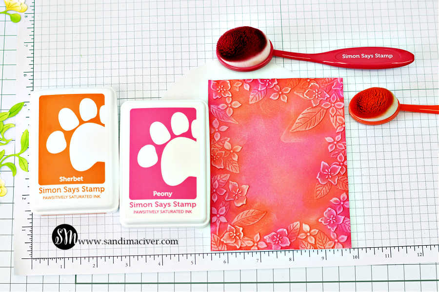 Ink-blended card background created with Simon Says Stamp Sherbert and Peony Pawsitively Saturated Inks using blending brushes. The background is embossed with the Fun Floral Frame Embossing Folder, highlighting floral and leaf patterns. Ink pads and brushes are shown on a gridded work surface alongside the finished colorful panel.