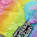 Handmade rainbow birthday card featuring a colorful ink-blended background embossed with seashells and sand dollars. White and gold paint splatters add texture and shimmer, while a bold black and white sentiment reads “Make your birthday a splash!” in the bottom right corner. Perfect for summer or beach-themed celebrations.