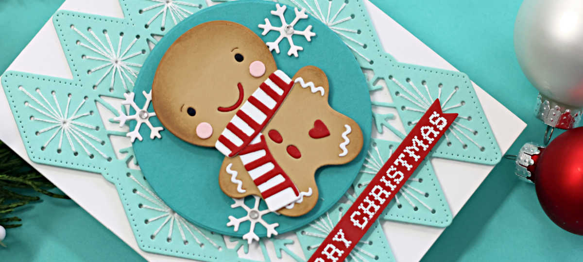 Handmade Christmas card featuring a die-cut gingerbread cookie with rosy cheeks and a red-and-white striped scarf, created using the Spellbinders July Stitched Club of the Month Frosted Stitch and the Caboodle Bonus Die Set of the Month Ginger Sip Die Set. The background showcases a teal stitched snowflake design layered on a white card base. A circular teal panel with white die-cut snowflakes frames the gingerbread figure. A red sentiment strip with a white heat-embossed "Merry Christmas" from the Stay Snuggly Clear Stamp and Die Set of the Month completes the festive design.
