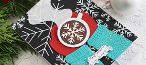 Handmade winter-themed card created using Spellbinders July Club dies, featuring layered die-cut elements including red mittens holding a hot chocolate mug with a snowflake design, textured turquoise sweater, and a sentiment reading "stay warm & sip happily" on a black background with bold white snowflake patterns.