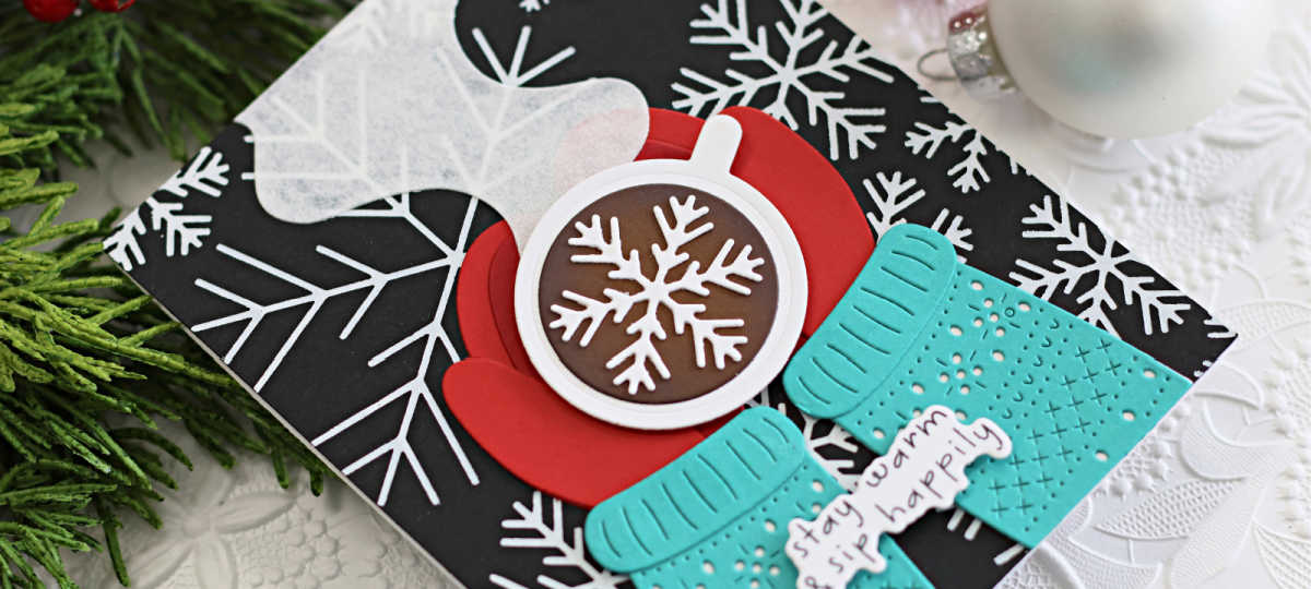 Handmade winter-themed card created using Spellbinders July Club dies, featuring layered die-cut elements including red mittens holding a hot chocolate mug with a snowflake design, textured turquoise sweater, and a sentiment reading "stay warm & sip happily" on a black background with bold white snowflake patterns.