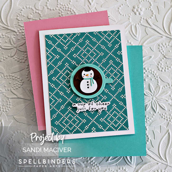 Handmade holiday card featuring Spellbinders BetterPress plate for the stitched background design, with a teal and white knit pattern. A die-cut cocoa mug with a snowman marshmallow sits in the center, paired with the stamped sentiment “a cup of cheer just for you.” Finished on a white card base