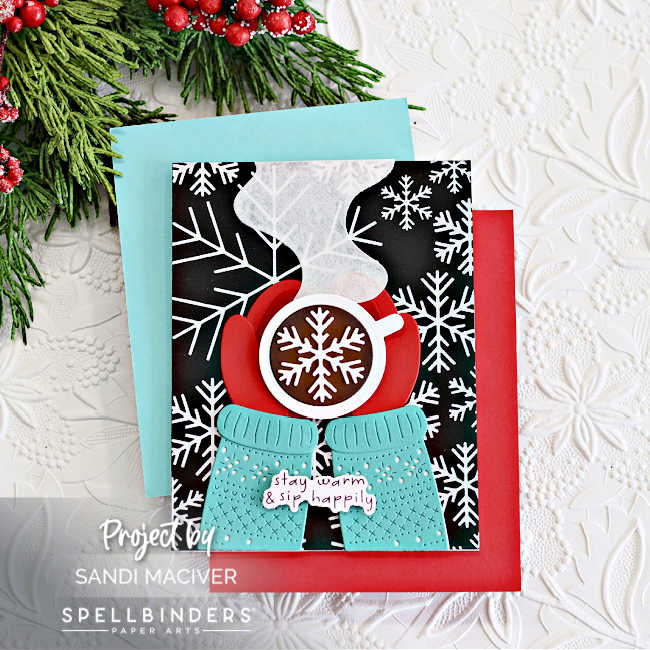Handmade winter-themed card created using Spellbinders July Club dies, featuring layered die-cut elements including red mittens holding a hot chocolate mug with a snowflake design, textured turquoise sweater, and a sentiment reading "stay warm & sip happily" on a black background with bold white snowflake patterns.