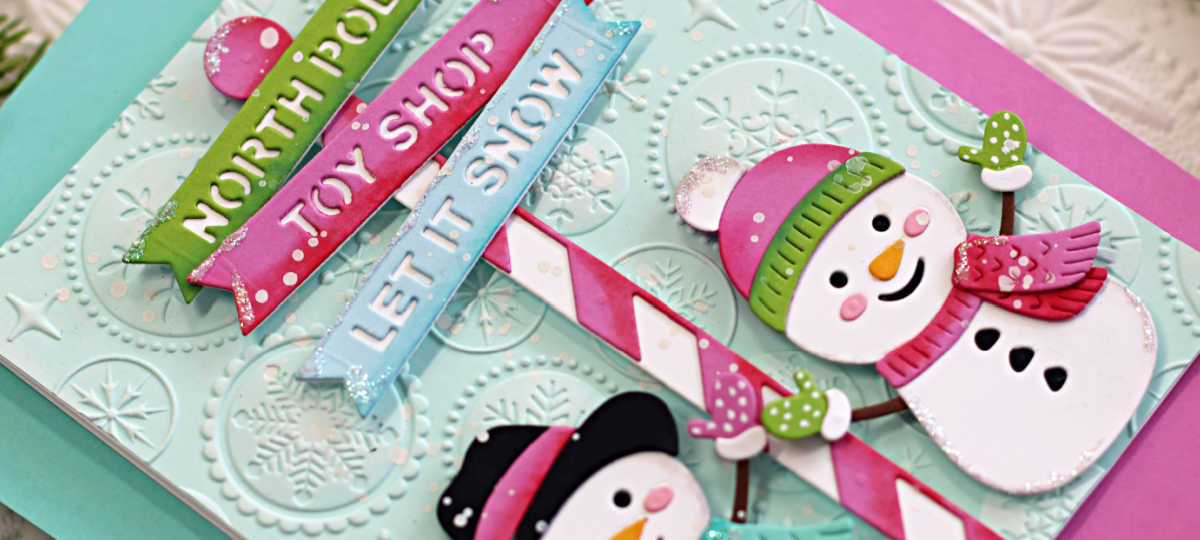 Close-up of a handmade Christmas card featuring Spellbinders North Pole Friends die cuts by Simon Hurley. The card showcases two smiling die-cut snowmen in pastel winter accessories, holding a red-and-white striped North Pole signpost with dimensional banners reading "NORTH POLE," "TOY SHOP," and "LET IT SNOW." The background is a soft aqua blue, embossed with the Starry Snowflakes 3D folder from the July 2025 Spellbinders release, adding festive texture and depth. Accents of glitter and white ink splatters enhance the whimsical, frosty holiday look.