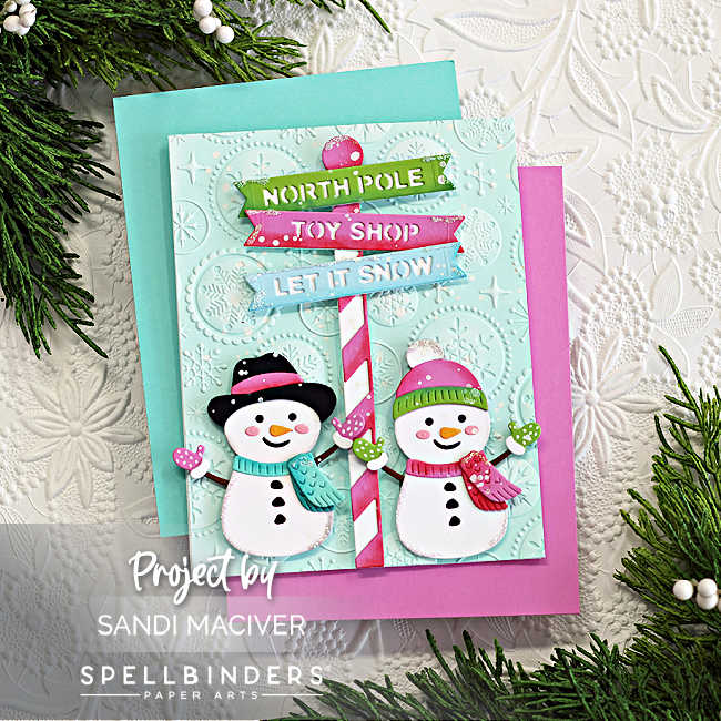 Handmade Christmas card featuring two cheerful die-cut snowmen created with the Spellbinders North Pole Friends die set by Simon Hurley. The snowmen wear colorful winter accessories and stand beside a red-and-white striped North Pole signpost with layered banners that read "NORTH POLE," "TOY SHOP," and "LET IT SNOW." The background is embossed with the Starry Snowflakes 3D folder on soft aqua cardstock, adding a festive and textured backdrop. The card is layered on pink and teal mats and styled with evergreen sprigs for a cozy holiday presentation.