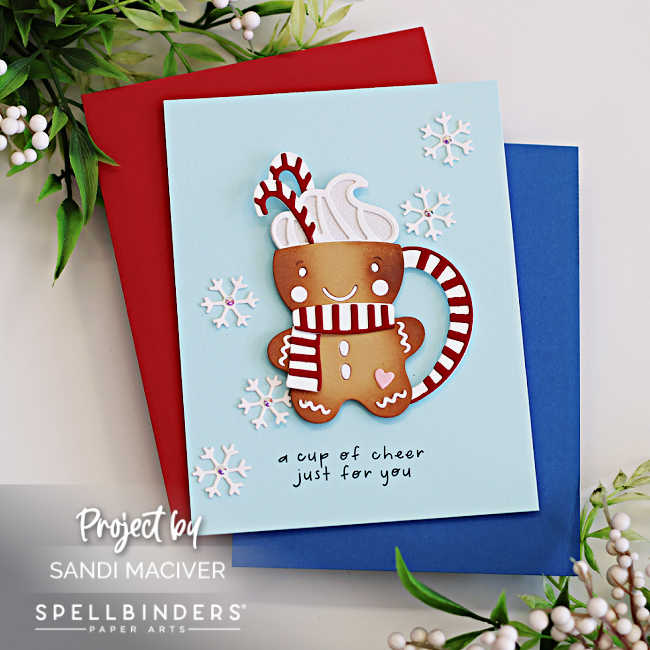 Handmade Christmas card featuring an adorable gingerbread man-shaped cocoa mug die cut with a red and white striped scarf, whipped cream, candy cane, and smiling face. The card has a light blue background decorated with white die-cut snowflakes and a stamped sentiment that reads “a cup of cheer just for you.” Created with Spellbinders Small Die of the Month by Sandi MacIver.