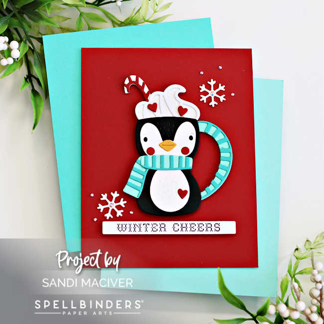 Handmade winter card featuring an adorable penguin-shaped cocoa mug die cut with whipped cream, candy cane, red hearts, and a turquoise striped scarf. The design sits on a bold red card front accented with white die-cut snowflakes and the sentiment “Winter Cheers.” Created with Spellbinders Small Die of the Month by Sandi MacIver.