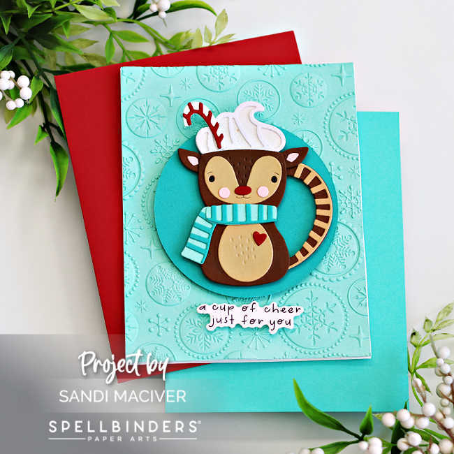 Handmade Christmas card featuring a cute reindeer-shaped cocoa mug die cut with whipped cream, candy cane, and a turquoise striped scarf on an embossed snowflake background. Sentiment reads “a cup of cheer just for you.” Created with Spellbinders Small Die of the Month by Sandi MacIver.
