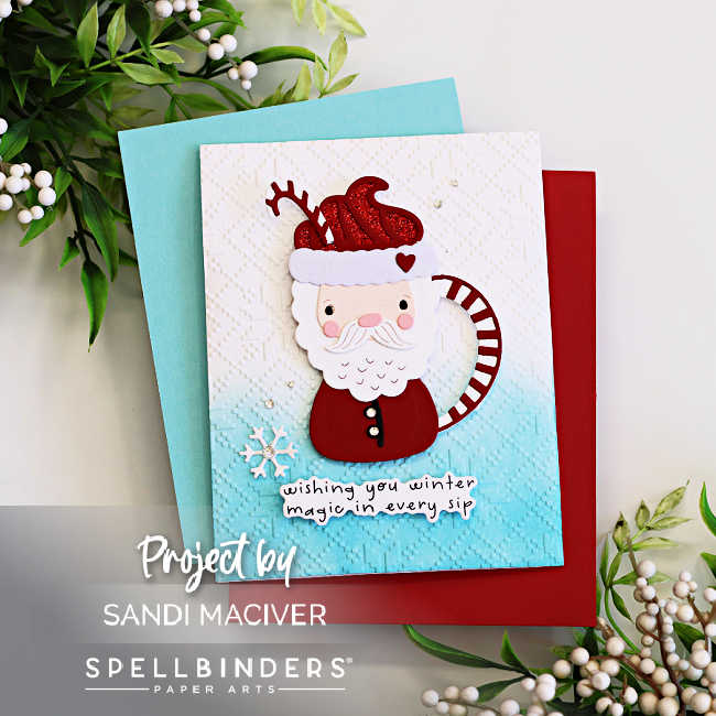  Christmas card featuring a Santa-shaped cocoa mug die cut with a red glitter hat, white beard, and candy cane handle on an embossed and ombre blue background. Embellished with a snowflake and sentiment reading “wishing you winter magic in every sip.” Created with Spellbinders Small Die of the Month by Sandi MacIver.