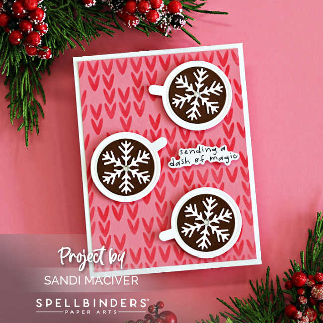Handmade Christmas card featuring three die-cut hot cocoa mugs with white snowflake toppers on a pink stenciled background created with the Spellbinders Warm and Woven stencil set, finished with the sentiment 'sending a dash of magic