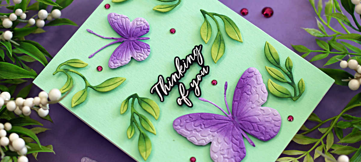 Handmade “Thinking of You” card featuring two embossed and die-cut butterflies from the Simon Says Stamp Confetti Butterflies embossing folders and dies, part of the Happy Camper Release. The butterflies are shaded in purple with textured wing details, surrounded by dimensional green leafy die cuts and pink gem accents, all arranged on a soft mint green card front. Card is styled on a purple background with greenery and white berry sprigs.