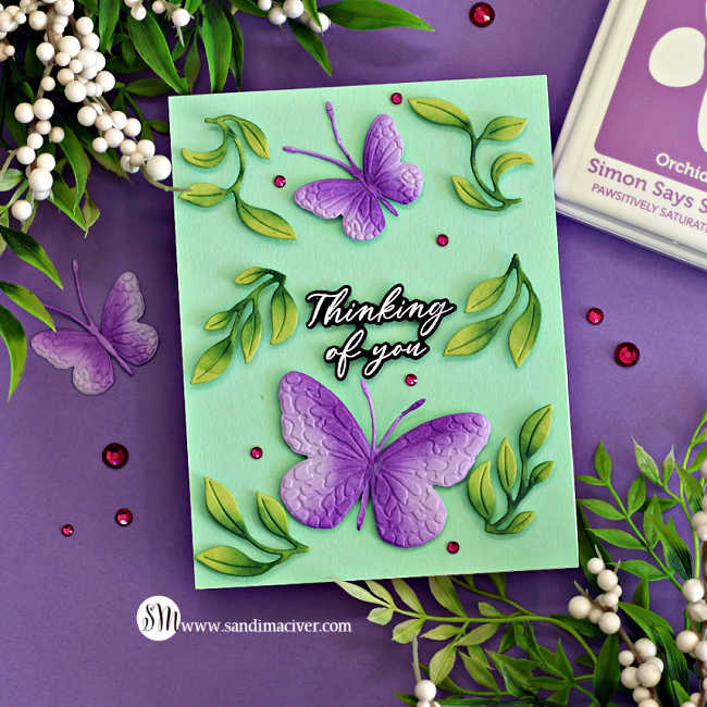 Handmade “Thinking of You” card featuring two embossed and die-cut butterflies from the Simon Says Stamp Confetti Butterflies embossing folders and dies, part of the Happy Camper Release. The butterflies are shaded in purple with textured wing details, surrounded by dimensional green leafy die cuts and pink gem accents, all arranged on a soft mint green card front. Card is styled on a purple background with greenery and white berry sprigs.