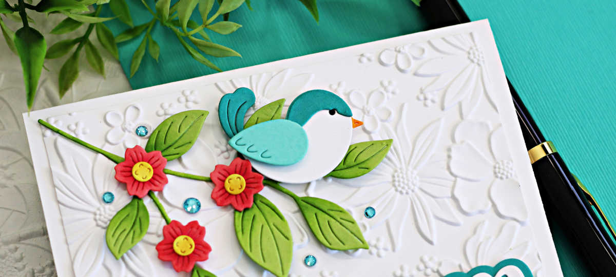 Handmade floral card featuring a die-cut teal and aqua bird perched on a leafy branch with red flowers and yellow centers, set against a white embossed floral background with blue gem embellishments – Simon Says Stamp ILYSM collection.