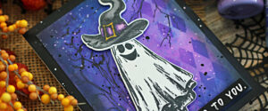 Handmade Halloween card featuring a ghost from the Simon Says Stamp Halloween Doodles stamp set and coordinating dies. The ghost wears a tall witch hat with a purple band and yellow buckle, layered over a vibrant background sprayed with Distress Oxide inks in Wilted Violet and Prize Ribbon. Black ink splatters and spooky tree silhouettes add texture, with a white heat-embossed sentiment strip in the bottom corner.