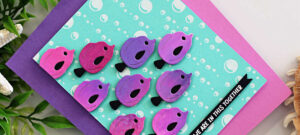 Handmade card featuring a school of whimsical die-cut fish created with the Spellbinders Glimmer of the Month and Deluxe Caboodle Bonus gift. The fish are arranged in neat rows across a turquoise background with Glimmer Hot Foiled white bubbles, blending from pink to purple hues with black tails. A small black banner in the lower right corner reads “WE ARE IN THIS TOGETHER” in white text. The design is mounted on a bright magenta card base, with a purple mat layer, evoking a playful and uplifting underwater scene.