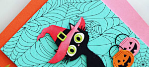 handmade Halloween card created with the Spellbinders Spooktacular release by Simon Hurley. The card features a black die-cut cat with large green eyes wearing a pink witch hat, layered over a teal BetterPress Spiderweb background. Pink and orange die0cut pumpkin treat baskets add a festive finishing touch.