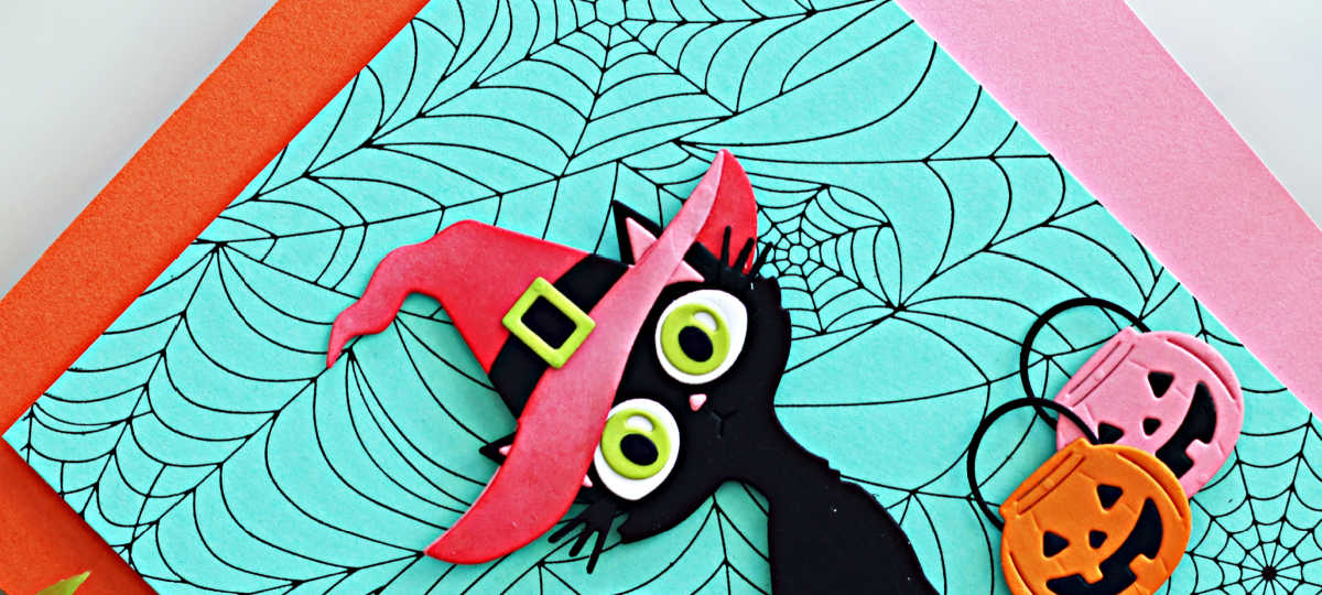 handmade Halloween card created with the Spellbinders Spooktacular release by Simon Hurley. The card features a black die-cut cat with large green eyes wearing a pink witch hat, layered over a teal BetterPress Spiderweb background. Pink and orange die0cut pumpkin treat baskets add a festive finishing touch.