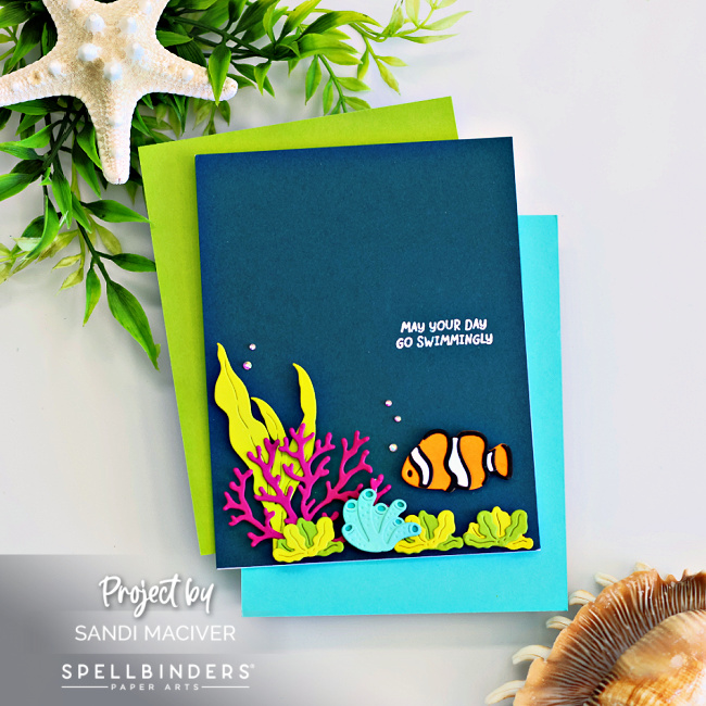 Handmade ocean-themed card created with the Spellbinders August Small Die of the Month Club Kit, Fish Friends Caboodle Bonus, and the Clear Stamp and Die Club. The deep teal card front features a white heat-embossed sentiment reading “May Your Day Go Swimmingly” near the top. At the bottom right, a vibrant underwater scene includes a bright orange and white clownfish, layered die-cut sea plants in lime green and aqua, and a bold pink coral branch. The card is accented with small iridescent gems to mimic air bubbles, and is photographed with seashells and greenery for a coastal vibe.