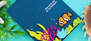 Handmade ocean-themed card created with the Spellbinders August Small Die of the Month Club Kit, Fish Friends Caboodle Bonus, and the Clear Stamp and Die Club. The deep teal card front features a white heat-embossed sentiment reading “May Your Day Go Swimmingly” near the top. At the bottom right, a vibrant underwater scene includes a bright orange and white clownfish, layered die-cut sea plants in lime green and aqua, and a bold pink coral branch. The card is accented with small iridescent gems to mimic air bubbles, and is photographed with seashells and greenery for a coastal vibe.