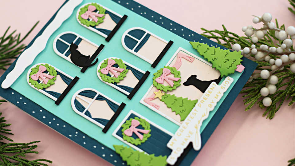 Handmade holiday card created with Spellbinders Yana’s City Holiday Collection dies, featuring a festive building facade with arched and square windows decorated with green wreaths and pink bows, playful black cat and dog silhouettes, layered Christmas trees, and a sentiment reading ‘My greatest present is having you by my side,’ set against a winter-themed background.