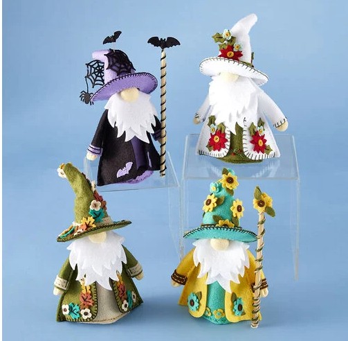 Four 3D Fergus felt gnomes created with the Spellbinders dies designed by Sandi MacIver, stitched in colorful felt with seasonal embellishment die sets. The gnomes feature detailed hats and robes decorated with flowers, leaves, and Halloween accents, each holding a staff and displayed against a light blue background.