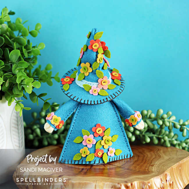 “Back view of handmade 3D felt Fergus the Gnome created with Spellbinders dies by Sandi MacIver. The gnome wears a bright teal coat and tall hat, both decorated with clusters of stitched felt flowers in pink, orange, and yellow with green leaves. Displayed on a wooden slice with greenery and a turquoise background