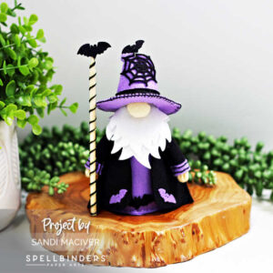 Handmade 3D felt Fergus the Gnome created with Spellbinders dies by Sandi MacIver. The gnome wears a purple hat with a stitched black spiderweb and black robe with purple accents, decorated with felt bats. He holds a striped staff topped with a black bat and is displayed on a wooden slice with greenery in the background.