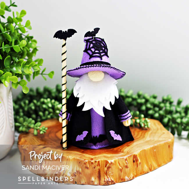 Handmade 3D felt Fergus the Gnome created with Spellbinders dies by Sandi MacIver. The gnome wears a purple hat with a stitched black spiderweb and black robe with purple accents, decorated with felt bats. He holds a striped staff topped with a black bat and is displayed on a wooden slice with greenery in the background.